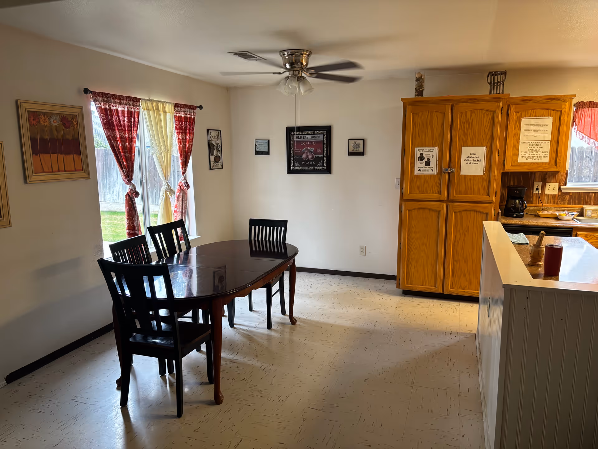A dining area with a dark wooden table and four matching chairs. The room has two windows with red and yellow curtains tied in the middle. On the right side, there is a wooden cabinet with notices posted on it, and a kitchen counter with a coffee maker and some items on it. The walls are decorated with framed pictures, and a ceiling fan with lights is mounted above the table.