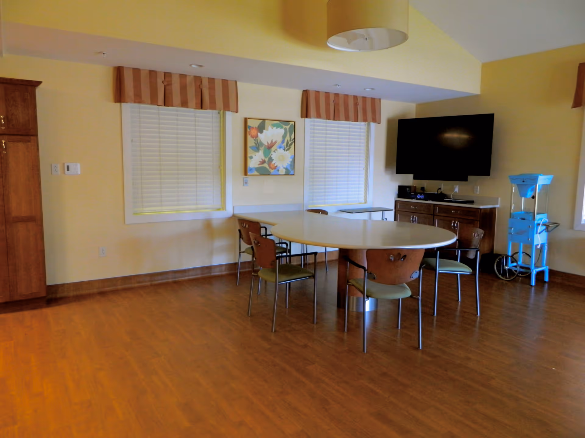 A room with wooden flooring and yellow walls featuring a large oval table surrounded by six chairs. Two windows with white blinds and striped valances are on the wall behind the table. A colorful floral painting hangs between the windows. A flat-screen TV is mounted on the wall above a wooden cabinet with drawers. A blue cart with wheels is positioned near the right window. A large ceiling light fixture hangs above the table.