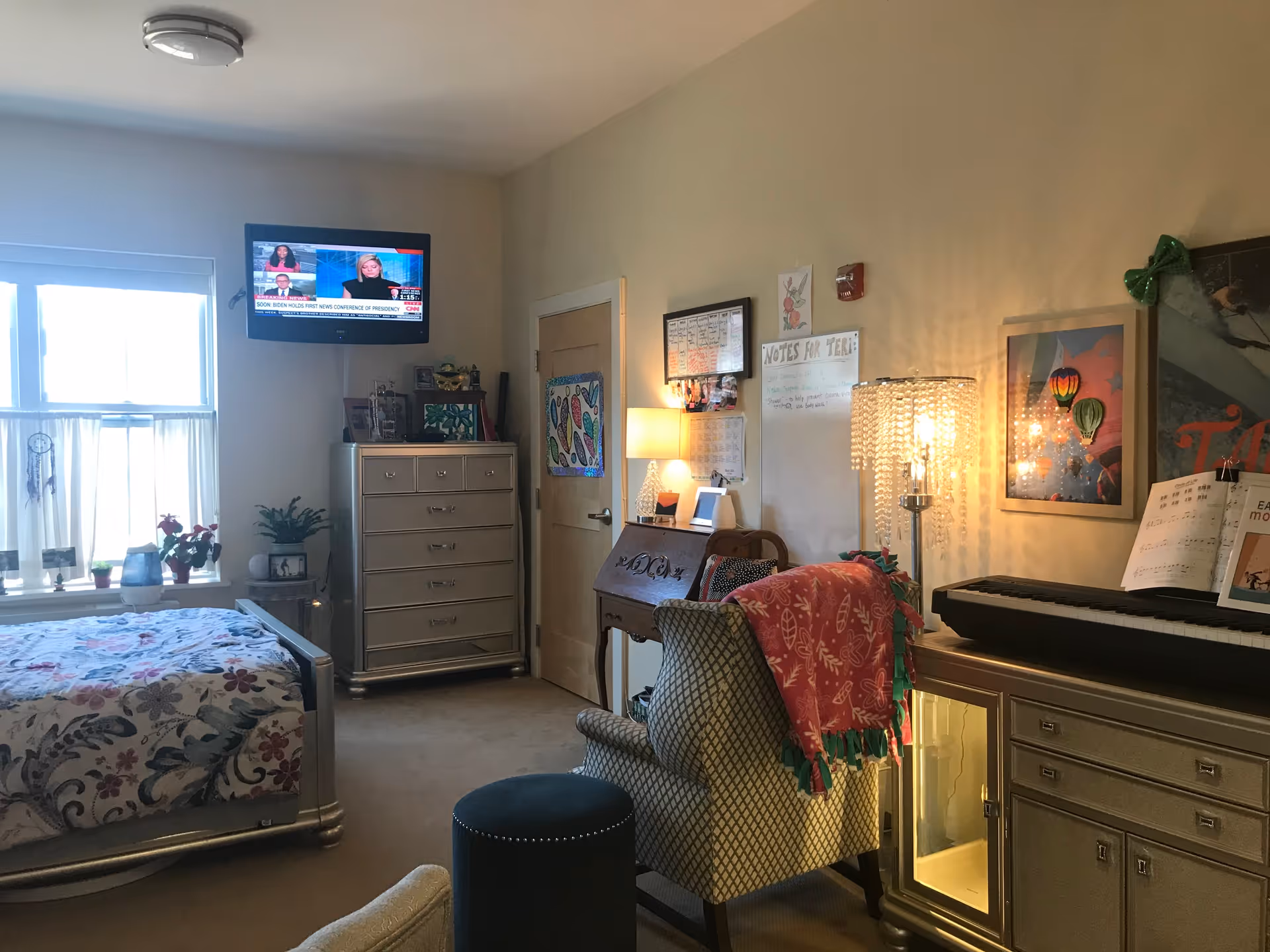 A cozy bedroom in a memory care facility featuring a bed with floral bedding near a window with sheer curtains. A dresser with drawers is positioned against the wall beneath a mounted TV showing a news channel. There is a comfortable armchair with a red blanket draped over it, a small round ottoman, a desk with a lamp, and a keyboard piano on a cabinet. The walls have framed pictures, a whiteboard with notes, and a door with colorful artwork.