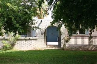 Exterior view of a brick building with a gated arched doorway, surrounded by greenery and trees, with a grassy lawn in the foreground.