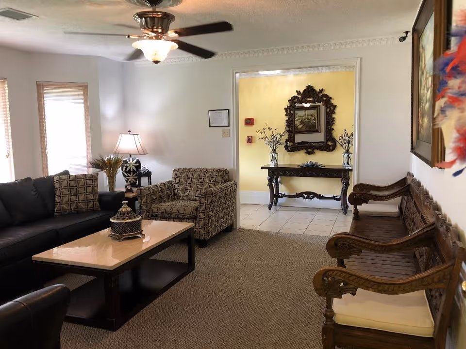 A cozy living room with a black sofa, patterned armchair, wooden coffee table with a decorative item, and a wooden bench with cushions. The room has a ceiling fan with a light, two windows with blinds, a table lamp, and a small side table with a plant. In the background, there is a yellow wall with a decorative mirror and a wooden console table with vases and flowers.