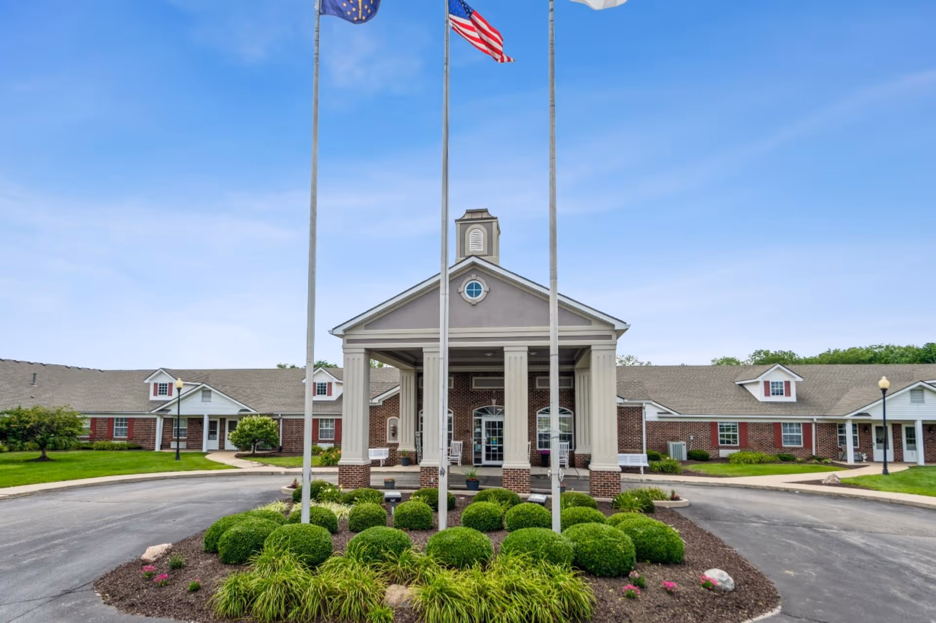 Front exterior view of Five Star Residences of Lafayette building with a covered entrance supported by columns, three flagpoles with flags, neatly trimmed bushes, and a circular driveway under a clear blue sky.