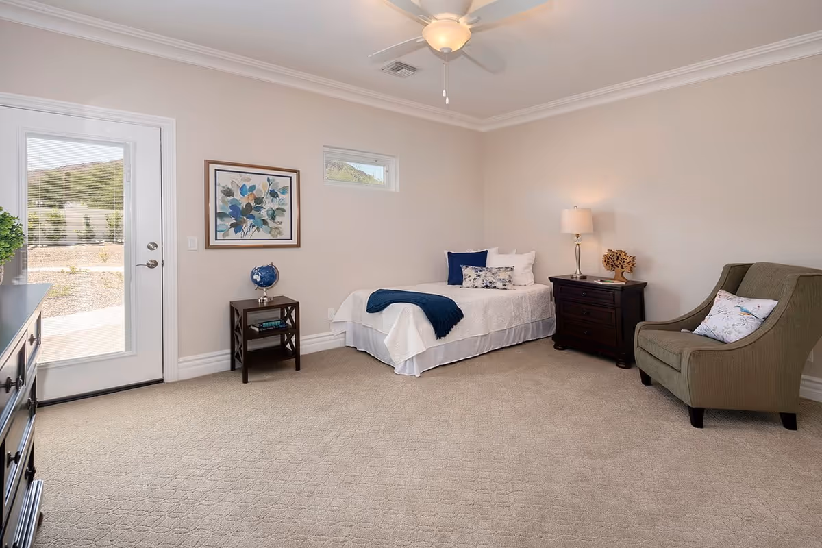 A bright and clean bedroom with beige walls and carpet. The room features a single bed with white bedding and blue accent pillows, a dark wooden nightstand with a lamp and decorative item, a matching small table with a globe and books, a comfortable green armchair with a floral pillow, a dresser, and a glass door leading outside with a view of a garden and hills.