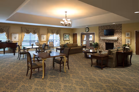 A spacious, well-lit common area with multiple wooden tables and chairs arranged on a patterned carpet. There are two sofas and a coffee table near a brick fireplace with a mounted TV above it. Large windows with draped curtains allow natural light to enter the room. A grand piano is visible in the corner, and framed artwork decorates the walls.