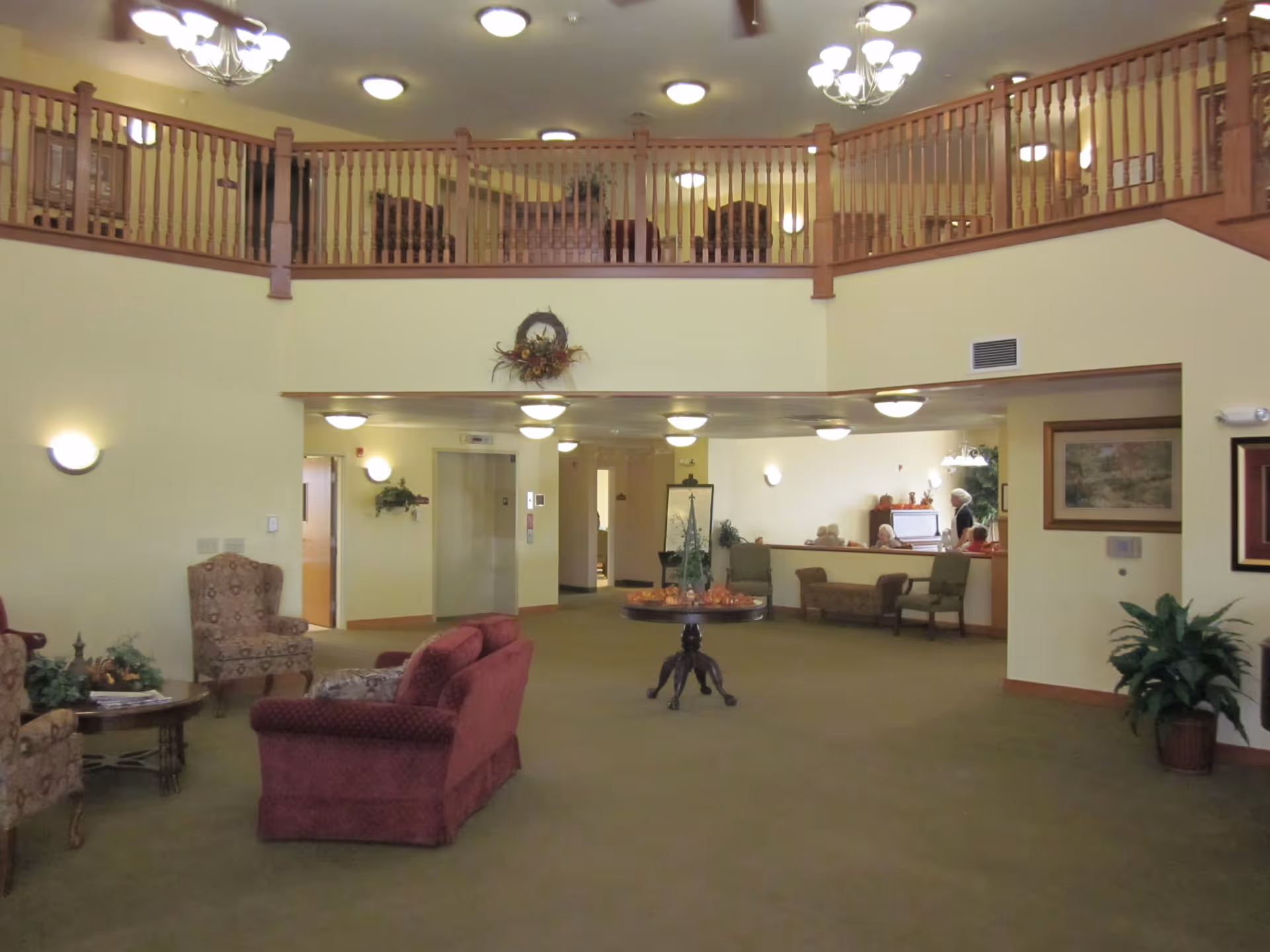 Spacious senior living facility common area with a high ceiling and wooden railing on the upper level. The room features comfortable upholstered chairs and sofas arranged around a small table, a round table with decorative items in the center, and an elevator in the background. There are several ceiling lights and wall sconces providing warm lighting, and a few people are visible in the background near a piano.