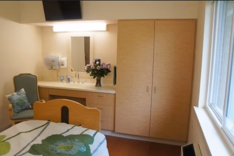 A senior living facility bedroom with a bed featuring a floral bedspread, a green cushioned chair with a decorative pillow, a built-in vanity with a mirror and a vase of flowers, and a large wooden wardrobe. There is a window letting in natural light on the right side.
