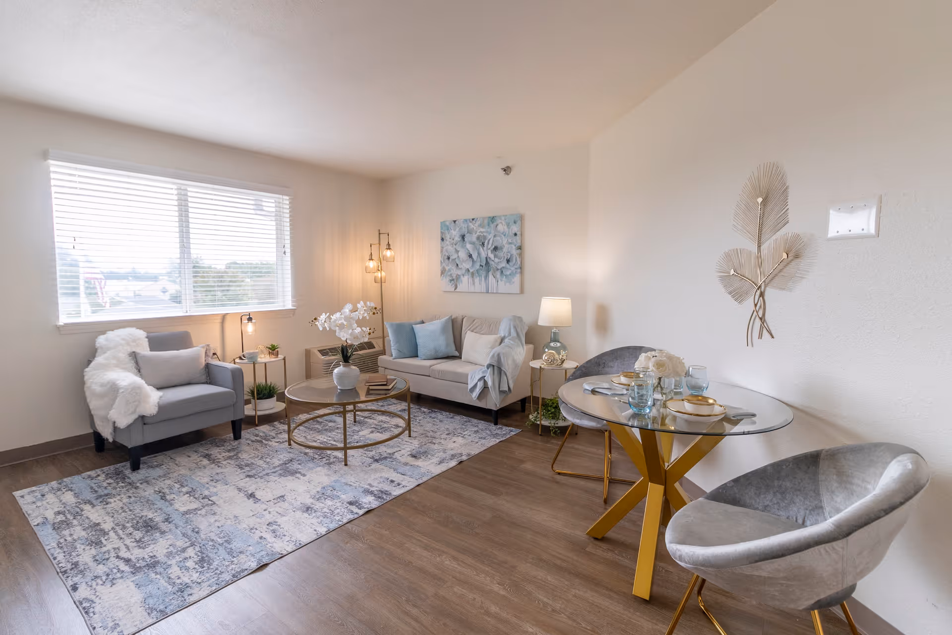 A bright and modern living room with a large window letting in natural light. The room features a gray armchair with a white throw, a beige sofa with blue and white cushions, a round glass coffee table with a white vase and flowers, and a blue patterned rug. There is a round glass dining table with gold legs set with plates, glasses, and cutlery, accompanied by two gray velvet chairs. The walls are decorated with a floral painting and a metal leaf wall art. The floor is wooden.