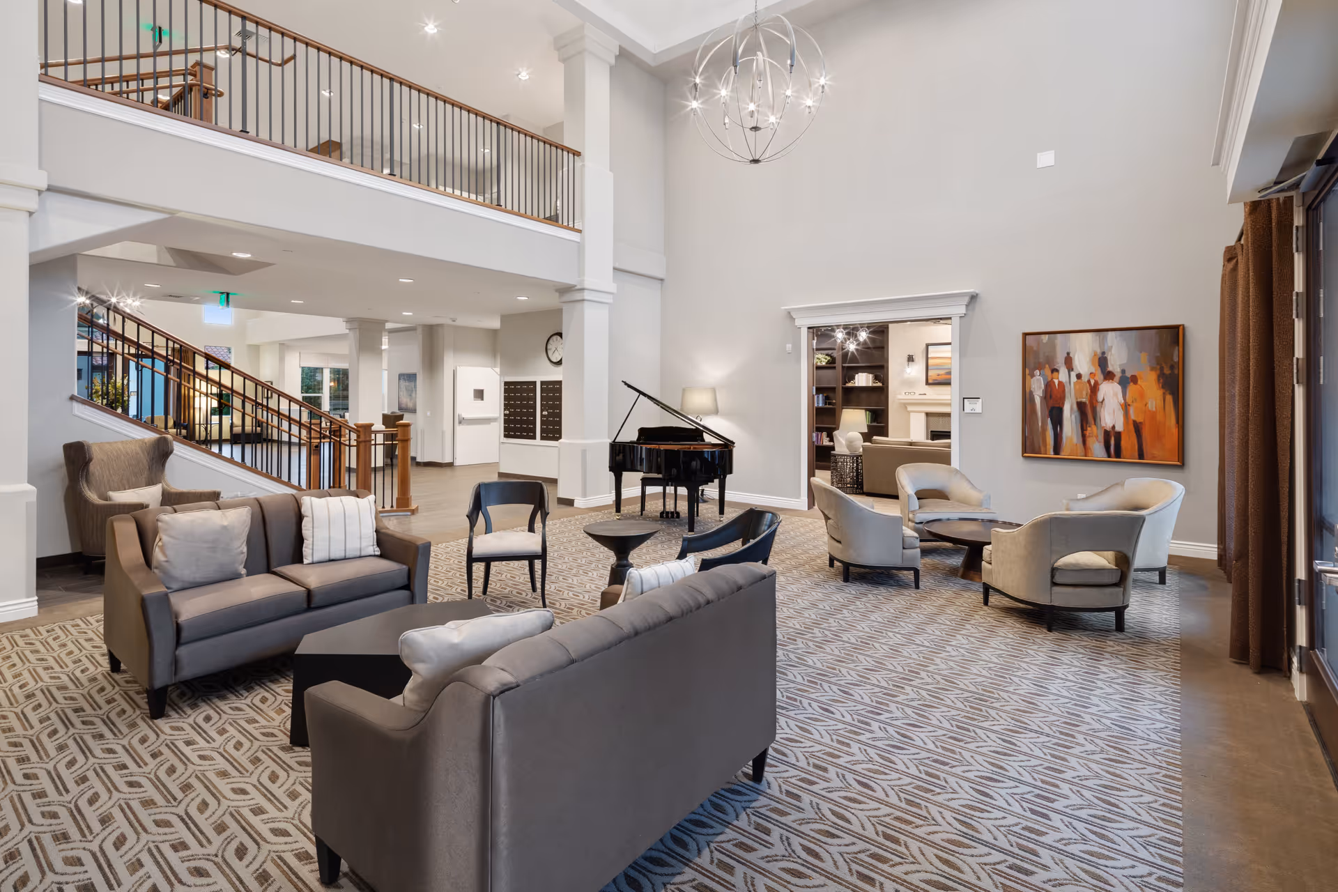 A spacious and well-lit living room area in a senior living facility featuring modern furniture including sofas, armchairs, and small tables arranged on a patterned carpet. A black grand piano is positioned near the center, with a staircase and balcony visible in the background. The walls are painted light gray, adorned with a colorful painting and a large modern chandelier hanging from the high ceiling.