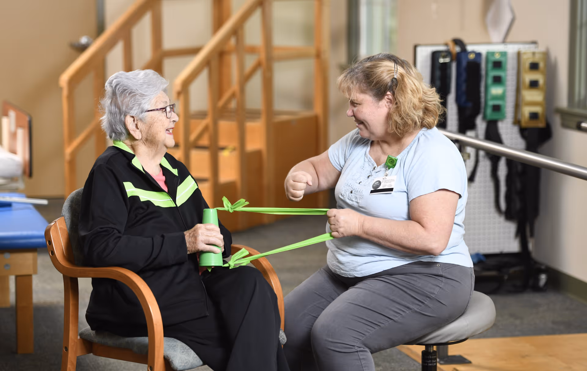 An elderly woman sitting in a chair exercises with a green resistance band held by a female caregiver or therapist in a rehabilitation or therapy room with exercise equipment in the background.