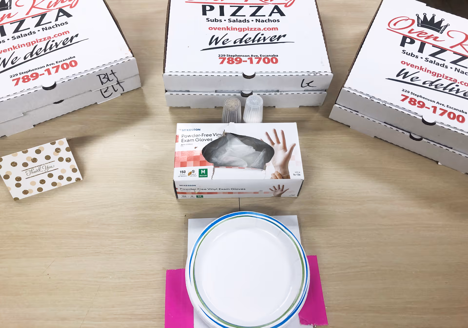 A wooden table with three stacked Oven King Pizza boxes, a box of powder-free vinyl exam gloves, salt and pepper shakers, a white paper plate with a blue and green rim on top of a pink napkin, and a small thank you card with gold polka dots.