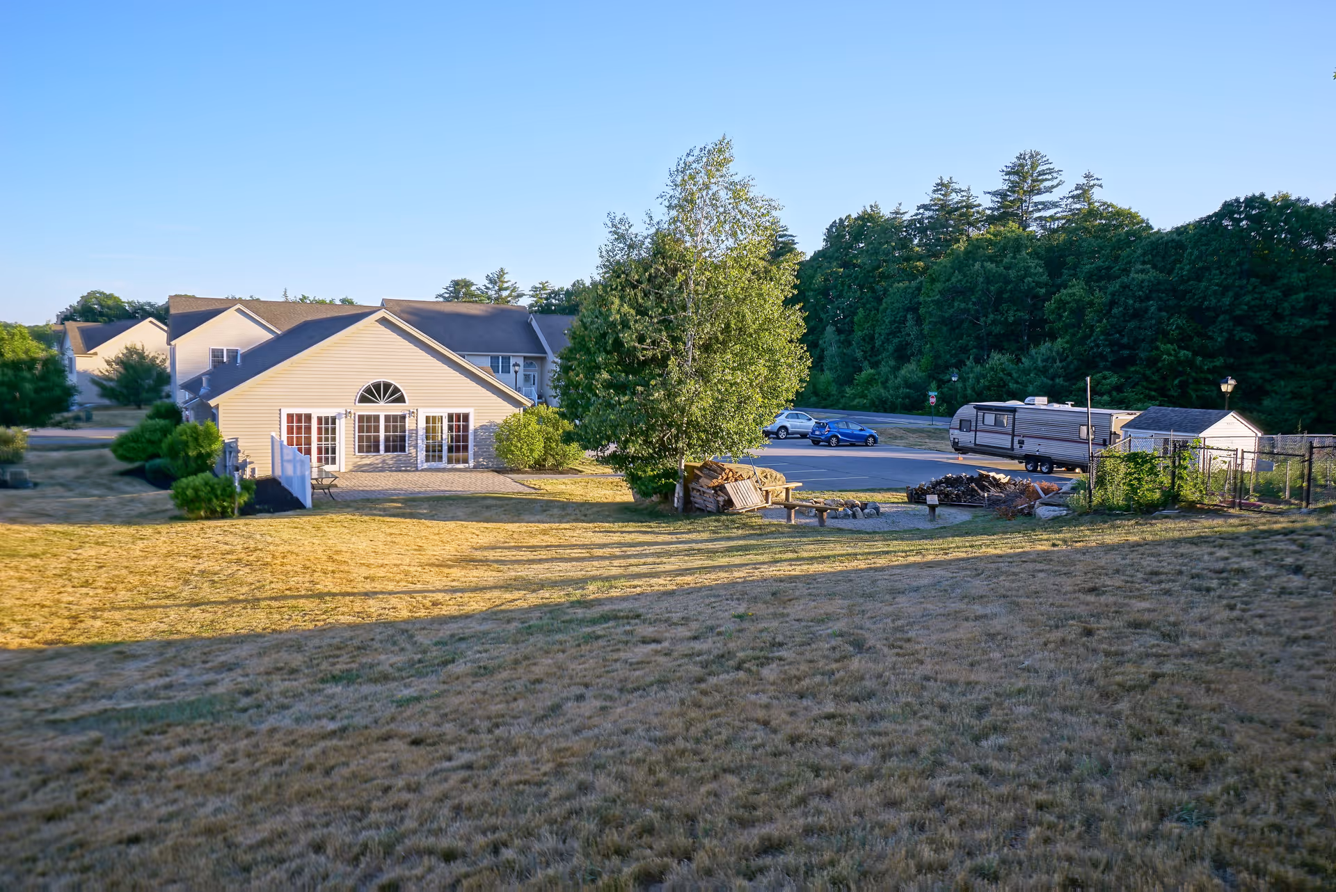 View of a residential area with a beige house featuring large windows and a patio, surrounded by trees and a grassy lawn. There is a parking area with a blue car and a camper trailer, and a small shed near the edge of the property. The scene is set under a clear blue sky.
