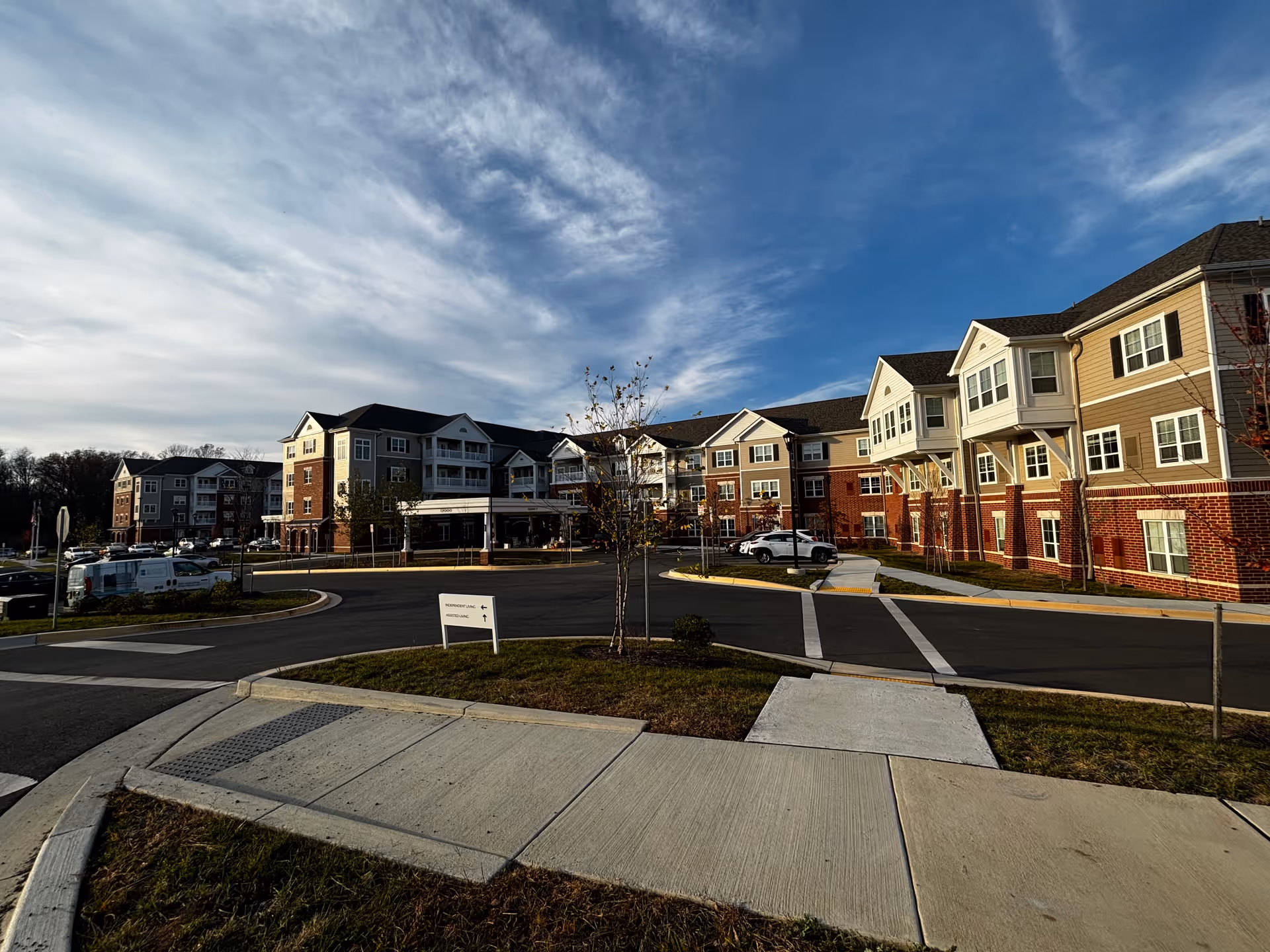 Front exterior of a multi-story senior living facility with a circular driveway, parked cars, and a blue sky.