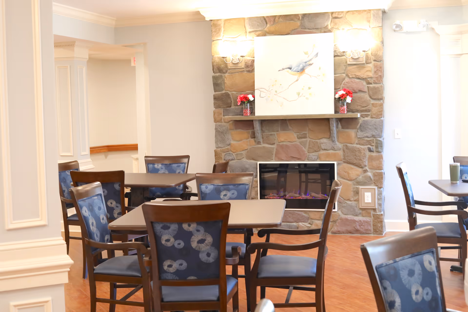 A cozy dining area with several wooden tables and chairs featuring blue patterned upholstery. The room has a stone fireplace with an electric fire and a painting of a bird above it. The walls are light-colored with decorative molding, and the floor is wooden.