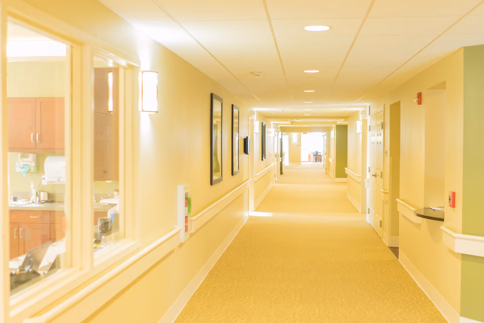 A long, well-lit hallway in a senior living facility with beige walls and carpet. The hallway features handrails on both sides, framed pictures on the left wall, and several doors along the right side. A window on the left reveals a room with wooden cabinets and a countertop.