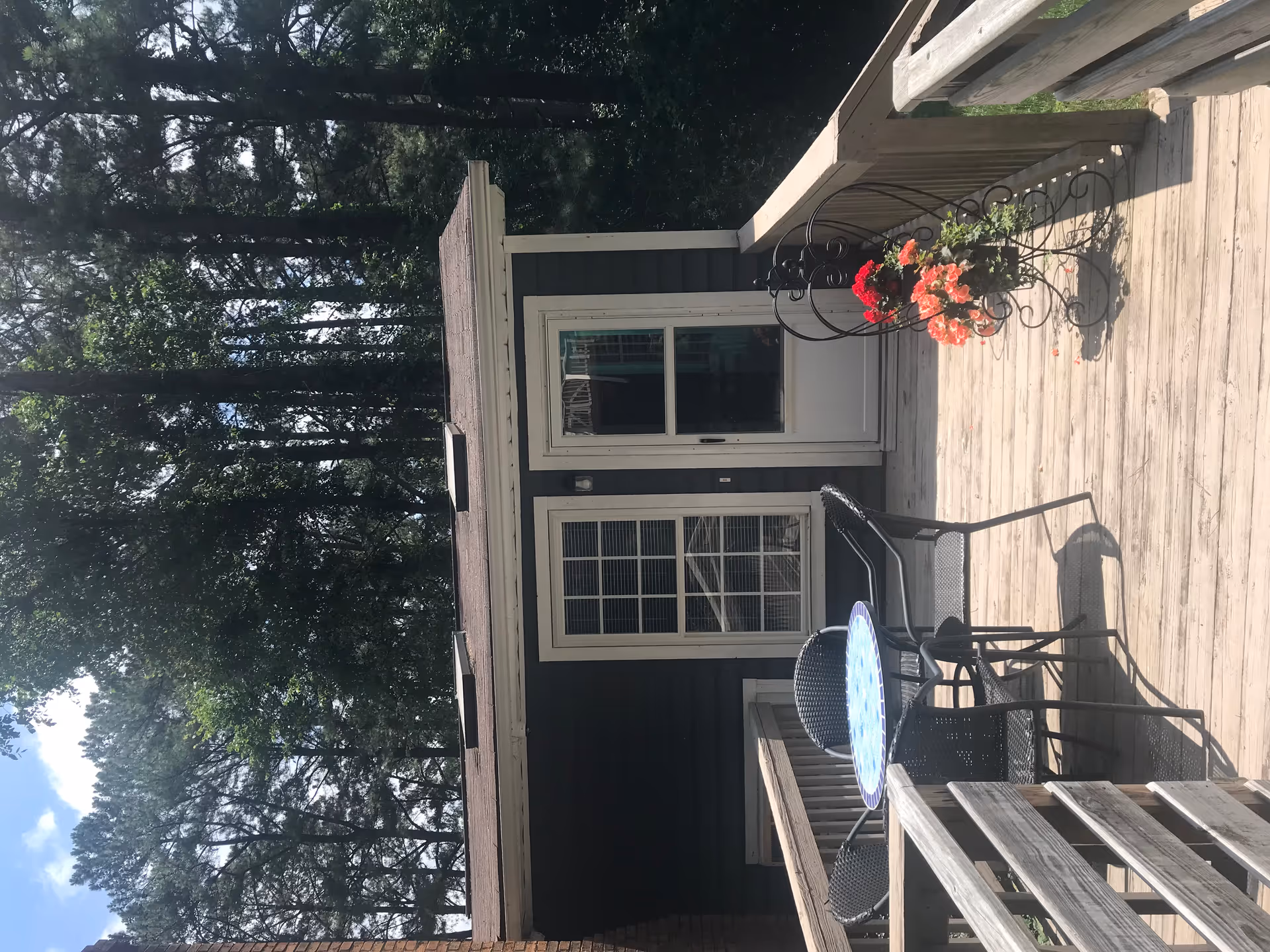 Outdoor wooden deck area with a small round table and four black chairs. There is a decorative black metal plant stand with red and pink flowers on the right side. The deck leads to a dark-colored building with a white-framed window and a white door. Tall trees and greenery are visible in the background.