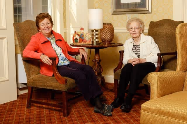 Two elderly women sitting in armchairs in a warmly decorated living room with patterned carpet and wallpaper. A round wooden table with a lamp and decorative items is between them.