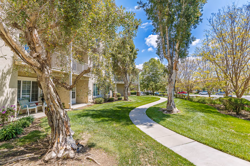 A sunny outdoor pathway winding through a landscaped garden area with green grass, trees, and shrubs. The pathway runs alongside a beige building with balconies and patio chairs visible. Several cars are parked in the background under a blue sky with scattered clouds.