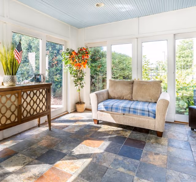 Sunlit sitting area with a beige loveseat topped by a blue plaid cushion, a decorative console table, potted plants and large windows overlooking greenery.