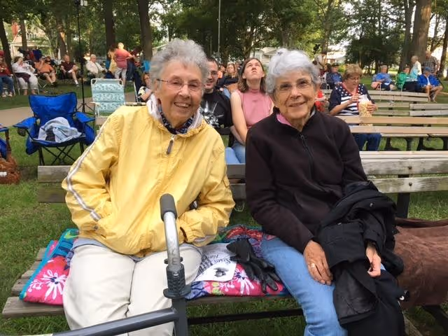 Two elderly women sitting on a wooden bench outdoors in a park-like setting. One woman is wearing a yellow jacket and glasses, holding a cane, and the other woman is wearing a black jacket and glasses. Behind them, several other people are seated on benches and folding chairs among trees and grass.
