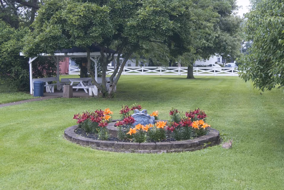 A well-maintained outdoor garden area with a circular flower bed containing orange and red flowers surrounding a decorative frog statue. In the background, there is a white wooden fence, several trees, and a covered picnic area with benches and a trash can.