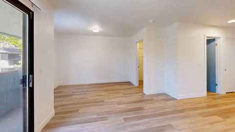 Empty room with light wood flooring, white walls, and a ceiling light fixture. There is a sliding glass door on the left side leading to an outdoor balcony. Two doorways are visible on the right side, one leading to a room with a blue wall and the other to a room with a wooden floor.