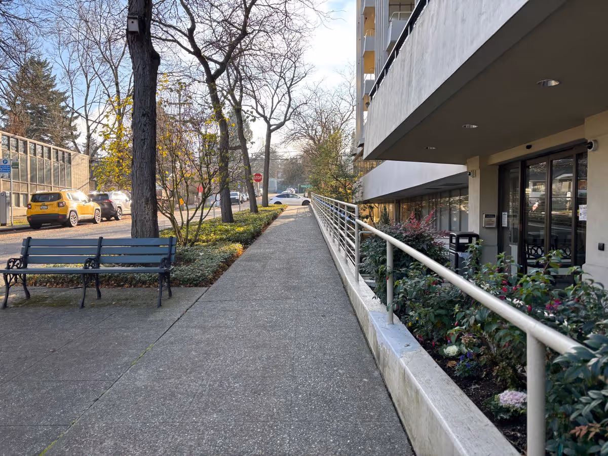 Concrete sidewalk and accessible ramp leading to the front entrance of a multi-story building, with benches, trees, and parked cars along the street.