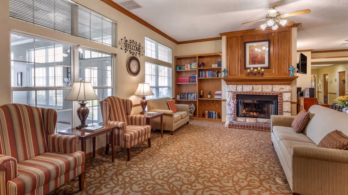 A cozy living room area in a senior living facility with striped armchairs, a beige sofa, two table lamps, a wall clock, a 'Welcome' sign, a wooden bookshelf filled with books, and a stone fireplace with a wooden mantle and framed artwork above it.