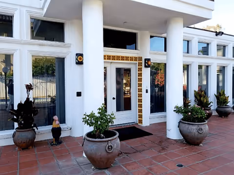 Front entrance of a white building with columns, decorative tile-framed door, and potted plants on a tiled walkway.