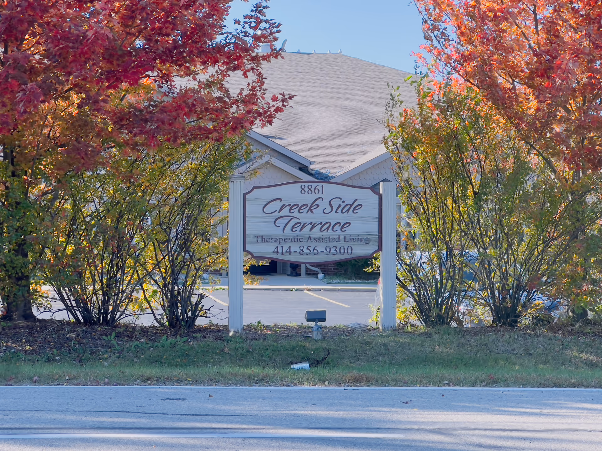 A wooden sign for Creek Side Terrace Therapeutic Assisted Living is positioned between two bushes with autumn-colored leaves. Behind the sign, there is a building with a gray roof and a parking lot. The sky is clear and blue.
