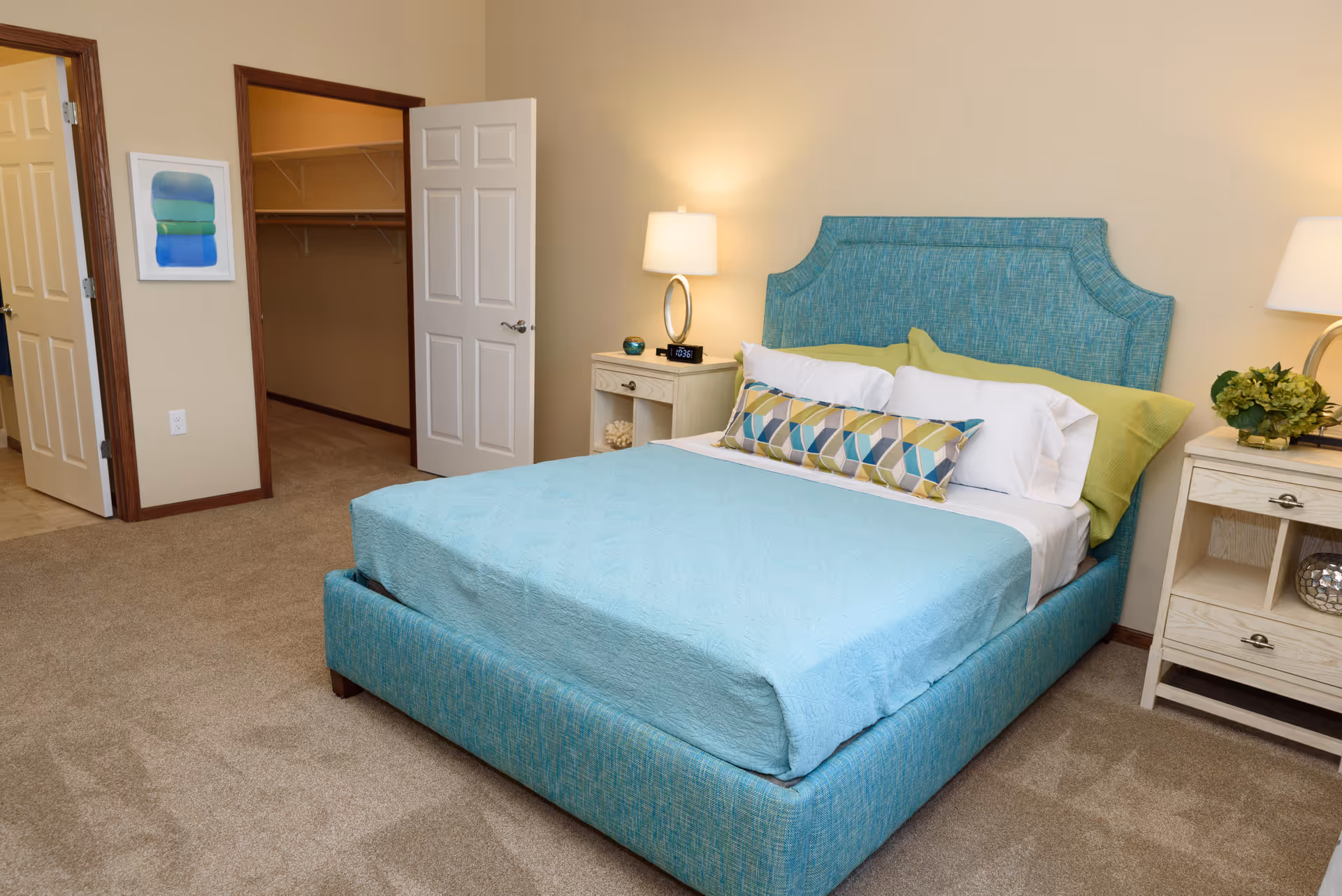 A neatly made bed with a turquoise upholstered headboard and matching bed frame, covered with a light blue quilt and decorative pillows. Two bedside tables with lamps flank the bed. An open door reveals a walk-in closet with shelving. The room has beige walls and carpeted floor.