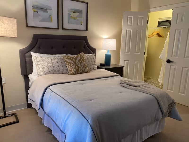 A neatly made bed with a gray upholstered headboard, white and patterned pillows, and a light gray blanket in a bedroom. There is a nightstand with a blue lamp and a small black object next to it. Two framed abstract paintings hang above the headboard. To the right, there is an open closet door showing hanging clothes and a bathrobe.
