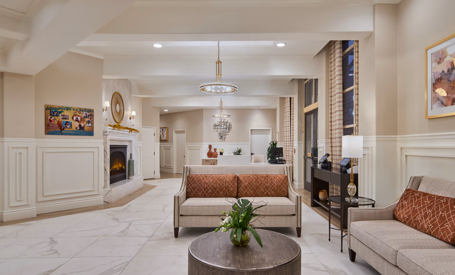Elegant senior living facility lobby with sofas, a coffee table with a plant, a fireplace, chandeliers, and a reception desk in the background.