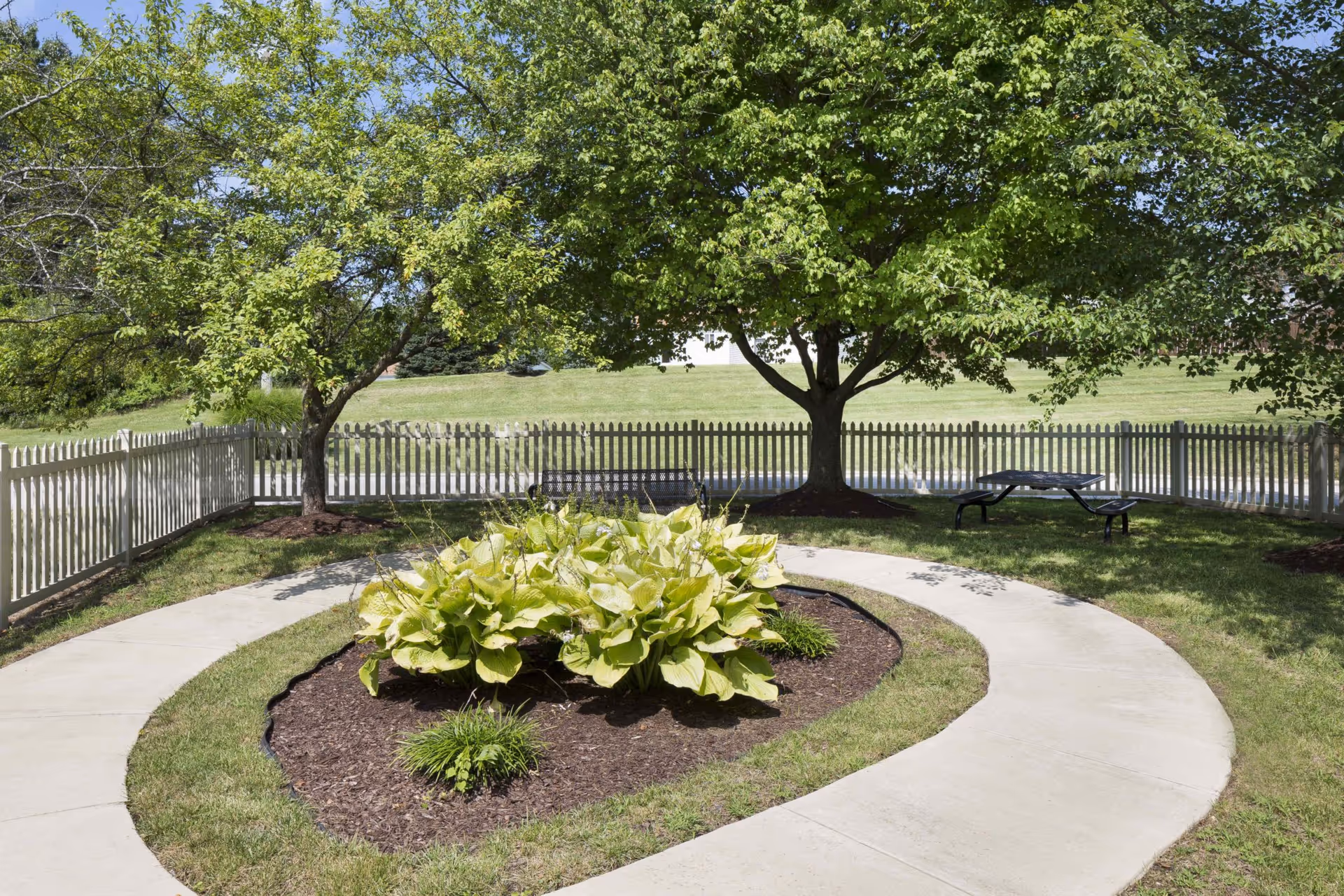 A peaceful outdoor garden area with a curved concrete walkway surrounding a mulched flower bed with green plants. Two large leafy trees provide shade, and a white picket fence encloses the space. A black metal picnic table is visible under one of the trees.