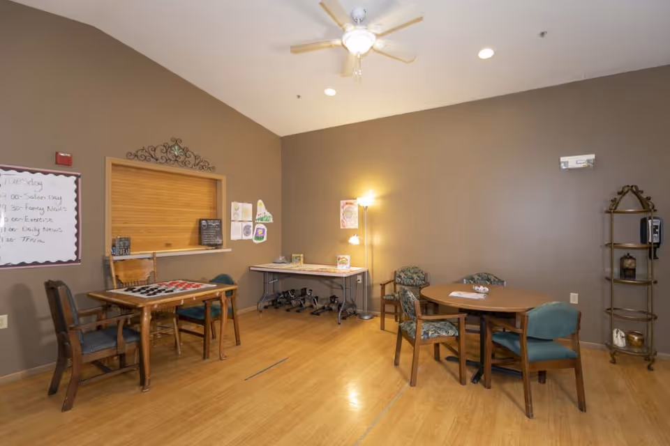 Interior community room with several wooden tables and chairs, a folding activity table, wall bulletin board, and a floor lamp.