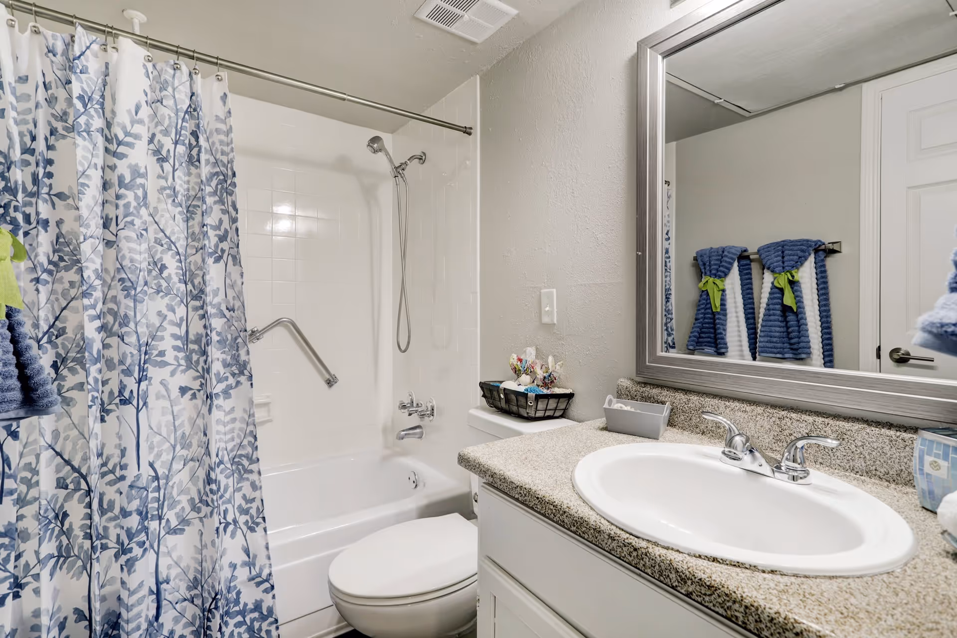 A clean bathroom featuring a white bathtub with a silver grab bar and a blue and white leafy patterned shower curtain. There is a white toilet next to the bathtub, and a granite countertop with a white sink and silver faucet. Above the sink is a large rectangular mirror with a silver frame. Blue towels with green accents hang on a towel rack reflected in the mirror.