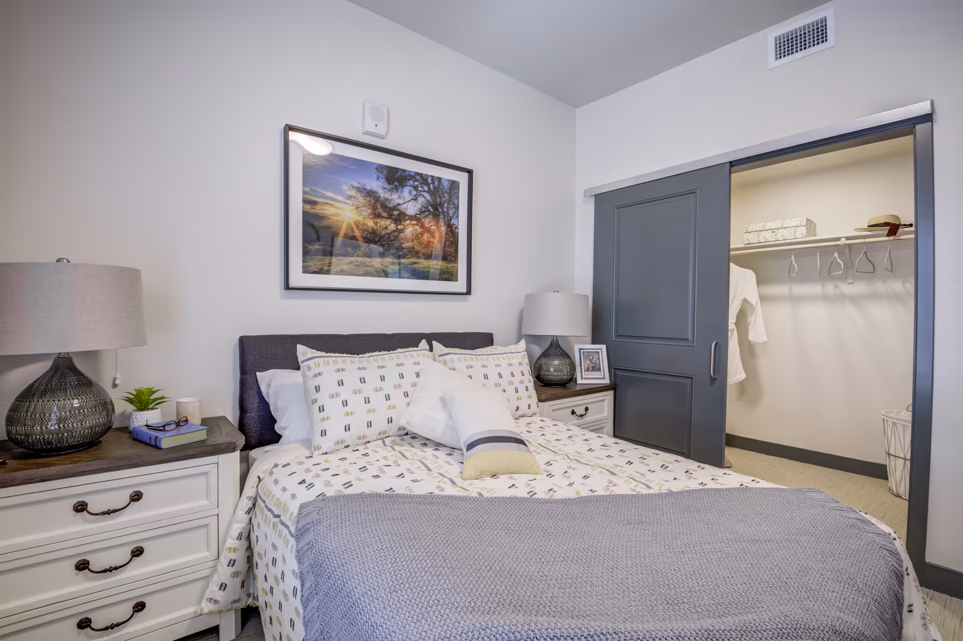 A neatly made bed with patterned bedding and multiple pillows in a bedroom. On either side of the bed are white nightstands with matching lamps, a small plant, books, and a framed photo. A large framed picture of a sunset over trees hangs on the wall above the bed. To the right, a sliding door is partially open, revealing a walk-in closet with hangers and storage boxes.