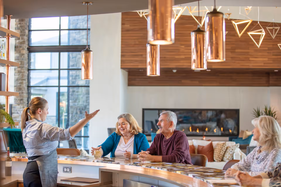 A group of three elderly people sitting at a curved bar counter in a modern lounge area, engaging with a female staff member standing behind the counter. The room features large windows, a stone wall, a fireplace, and contemporary pendant lighting.