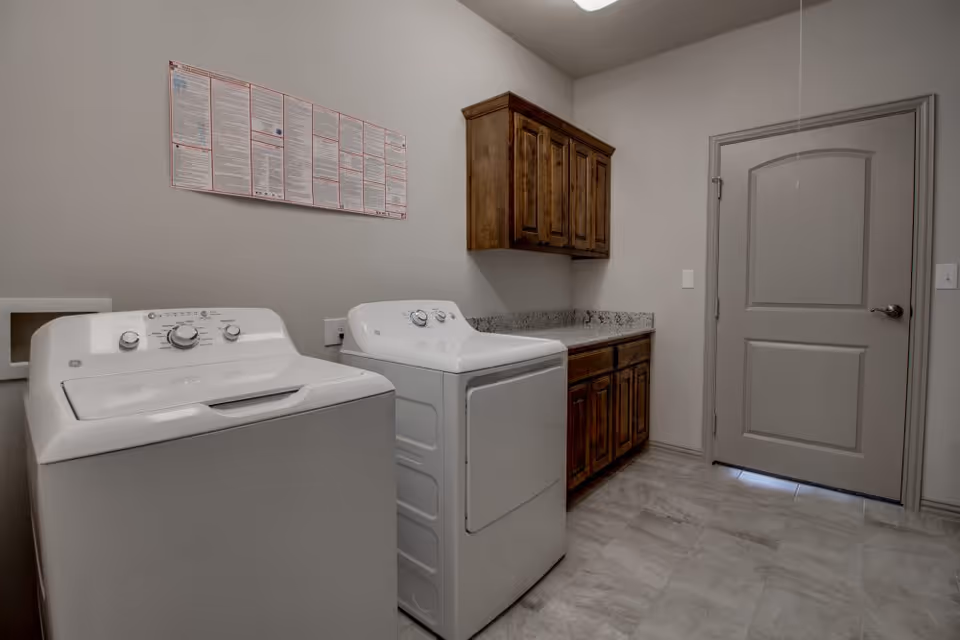 Laundry room with a white washing machine and dryer side by side, wooden cabinets above and below a granite countertop, a closed gray door, and a notice board on the wall.