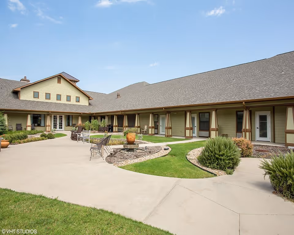 Outdoor courtyard area of Stonefield Assisted Living and Memory Care featuring a paved walkway, green grass, shrubs, potted plants, and several chairs and benches arranged for seating. The building surrounding the courtyard has a beige exterior with brown trim and multiple doors and windows under a sloped roof.