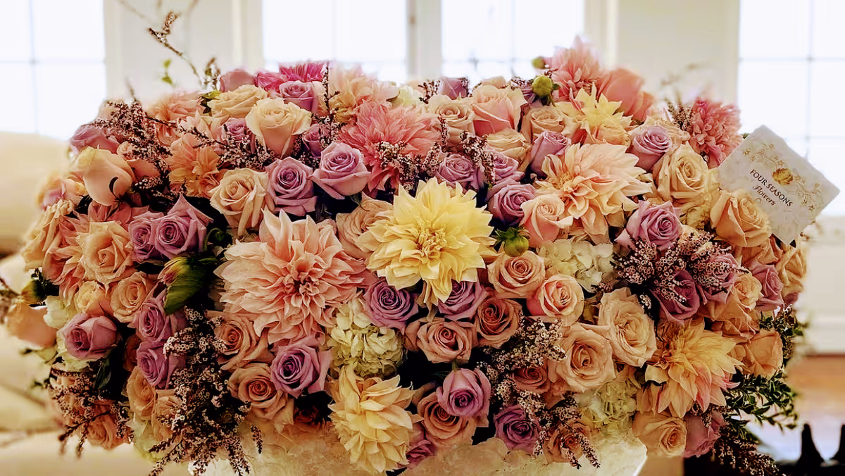 A large, colorful bouquet of flowers featuring pink, peach, purple, and cream roses along with other flowers arranged in a decorative display, set indoors with a softly lit background.