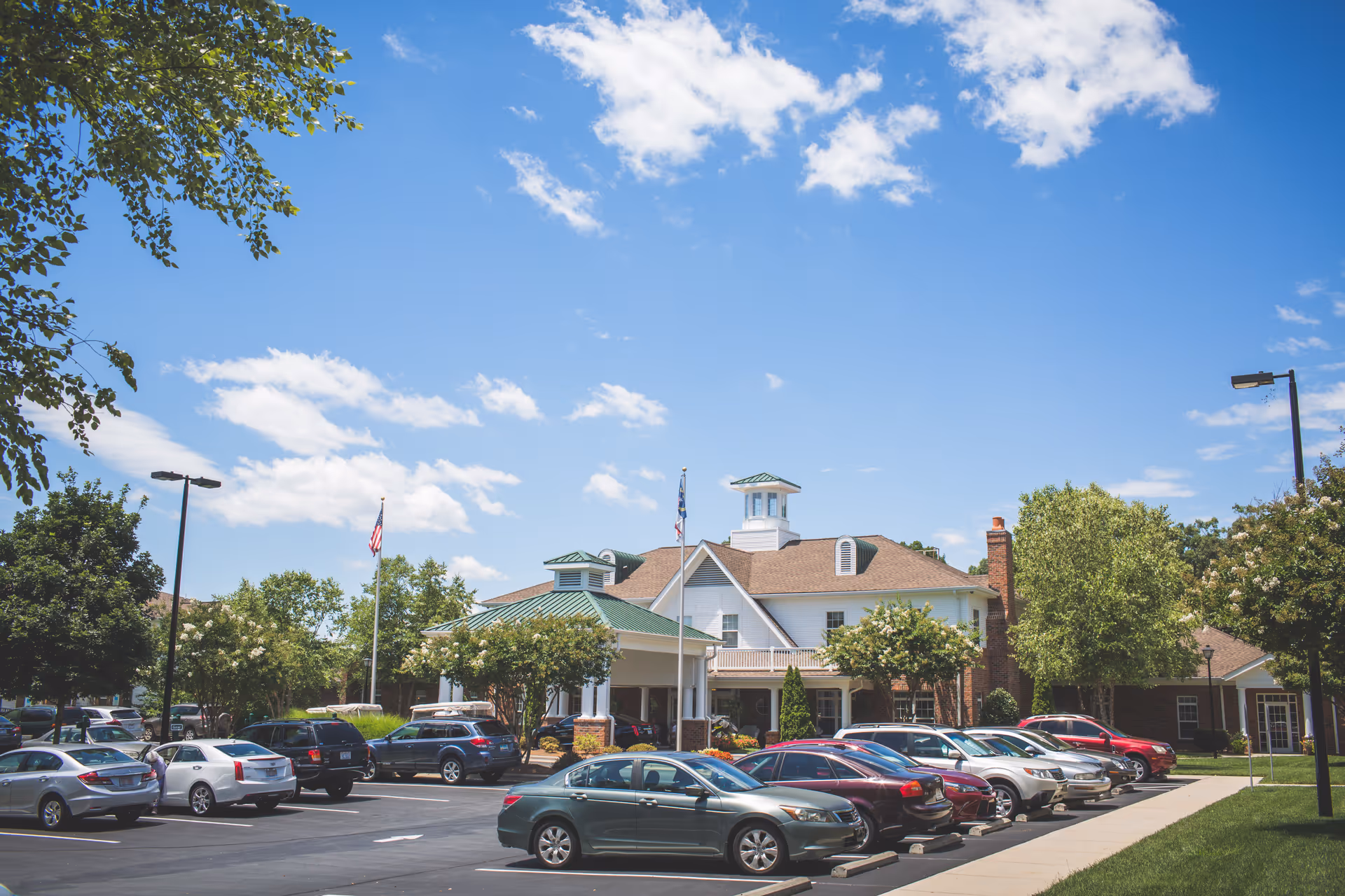 Exterior view of a senior living facility named Homestead Hills with a parking lot in front filled with cars, surrounded by trees under a blue sky with some clouds.