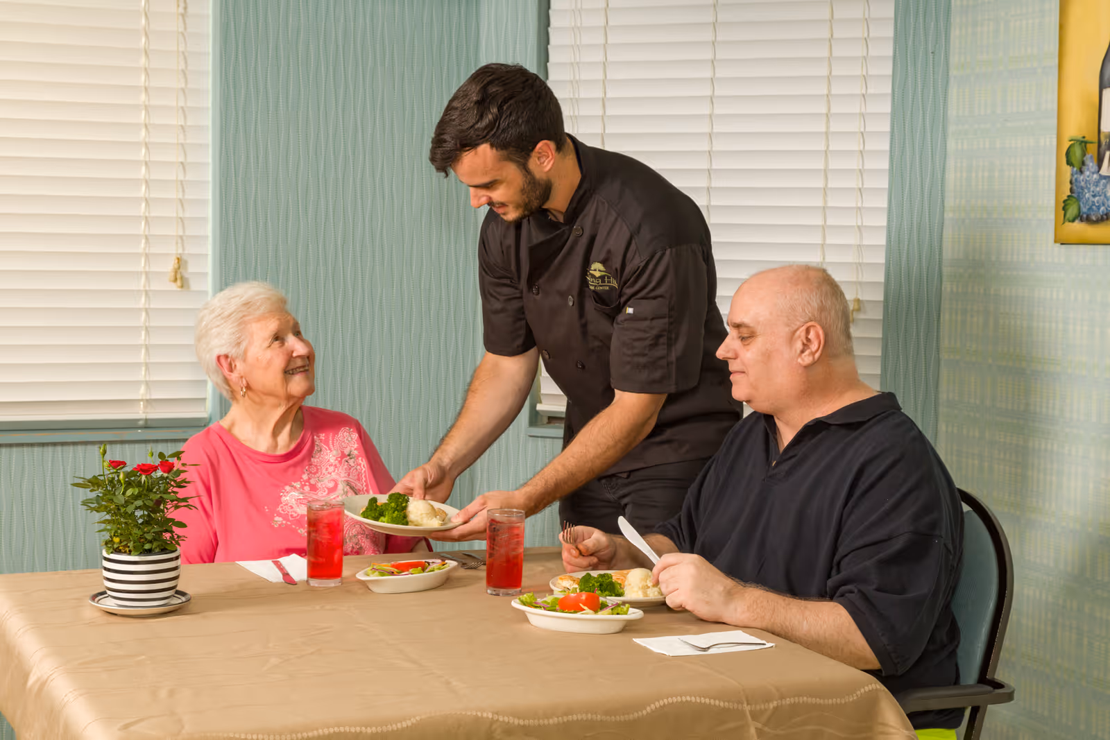 A staff member serves plates of food to two older adults seated at a dining table.