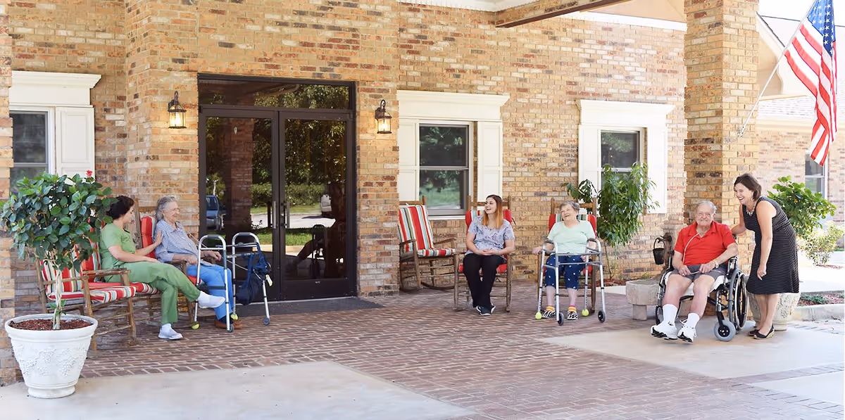 A group of elderly residents and caregivers sitting and interacting outside a brick building with large windows and an American flag. Some residents are seated in wheelchairs and using walkers, while others sit on chairs with red and white striped cushions. There are potted plants on either side of the entrance.
