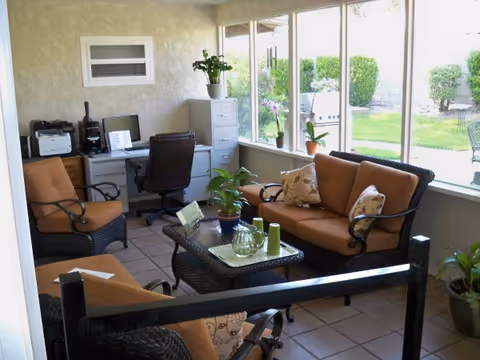 A cozy indoor sitting area with wicker furniture including a sofa and two armchairs with orange cushions and decorative pillows. A glass-top coffee table with plants and decorative items is in the center. A desk with a computer, printer, and office chair is against the wall. Large windows provide a view of a green outdoor garden with bushes and a barbecue grill.