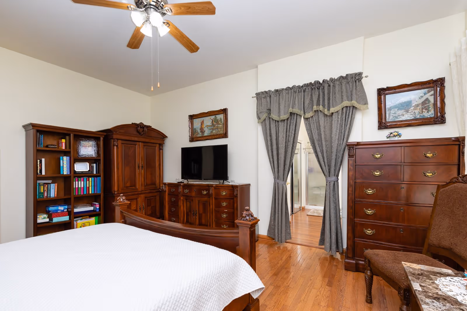 A bedroom with wooden furniture including a bed with a white quilt, a bookshelf filled with books and items, a large dresser with a flat-screen TV on top, and a tall chest of drawers. The room has hardwood floors, a ceiling fan with lights, and two framed paintings on the walls. Gray curtains hang over a doorway leading to a bathroom with a glass shower enclosure.