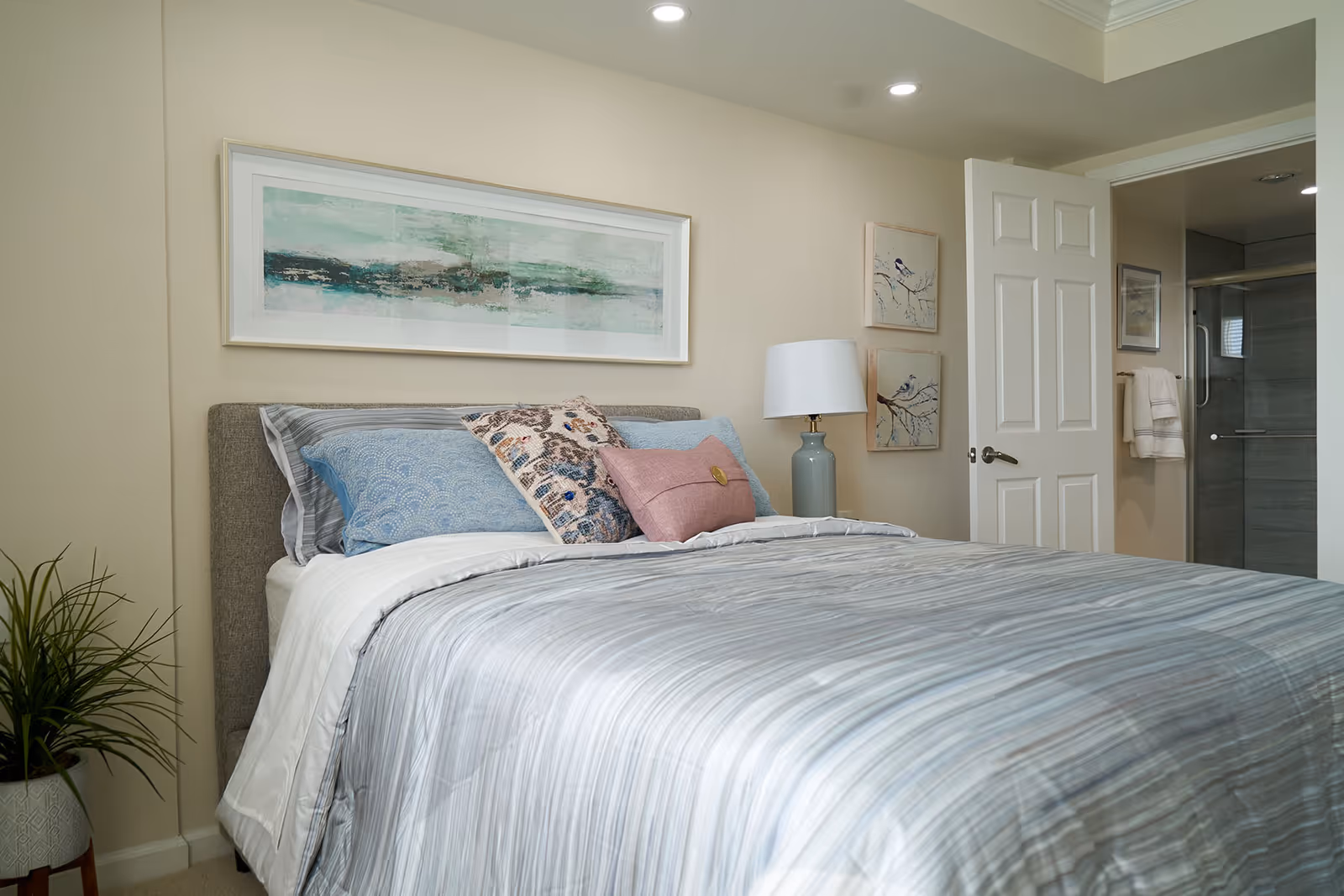 A neatly made bed with a gray upholstered headboard, striped bedding, and several decorative pillows in blue, pink, and patterned designs. Above the bed hangs a large abstract painting in shades of green and blue. To the right of the bed is a nightstand with a blue lamp and two framed bird illustrations on the wall. An open door reveals a bathroom with a glass shower and towels hanging on the wall. A potted plant is visible on the left side of the bed.