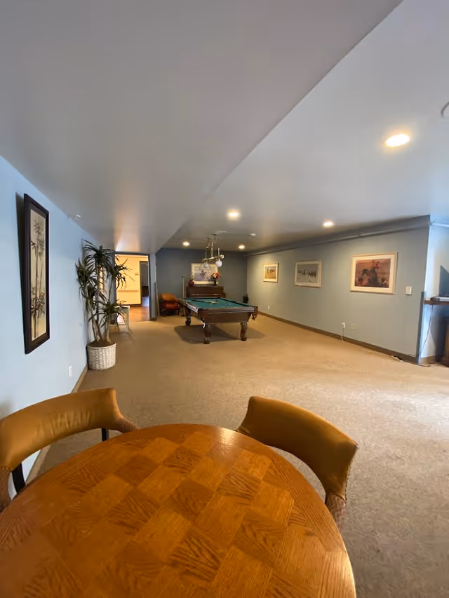 A spacious recreational room with beige carpet and light blue walls. In the foreground, there is a round wooden table with two brown chairs. In the middle of the room, there is a pool table with overhead lighting. The walls are decorated with framed artwork, and there is a tall potted plant near the left wall. The room is well-lit with recessed ceiling lights.