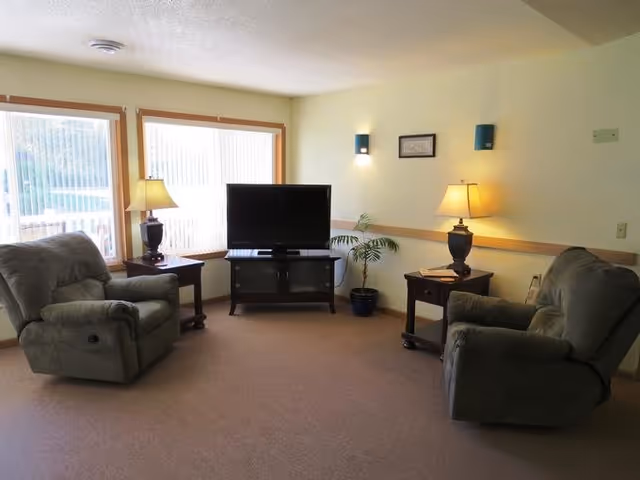 Small living room with two recliners facing a TV on a stand between windows, with side tables, lamps and a potted plant.