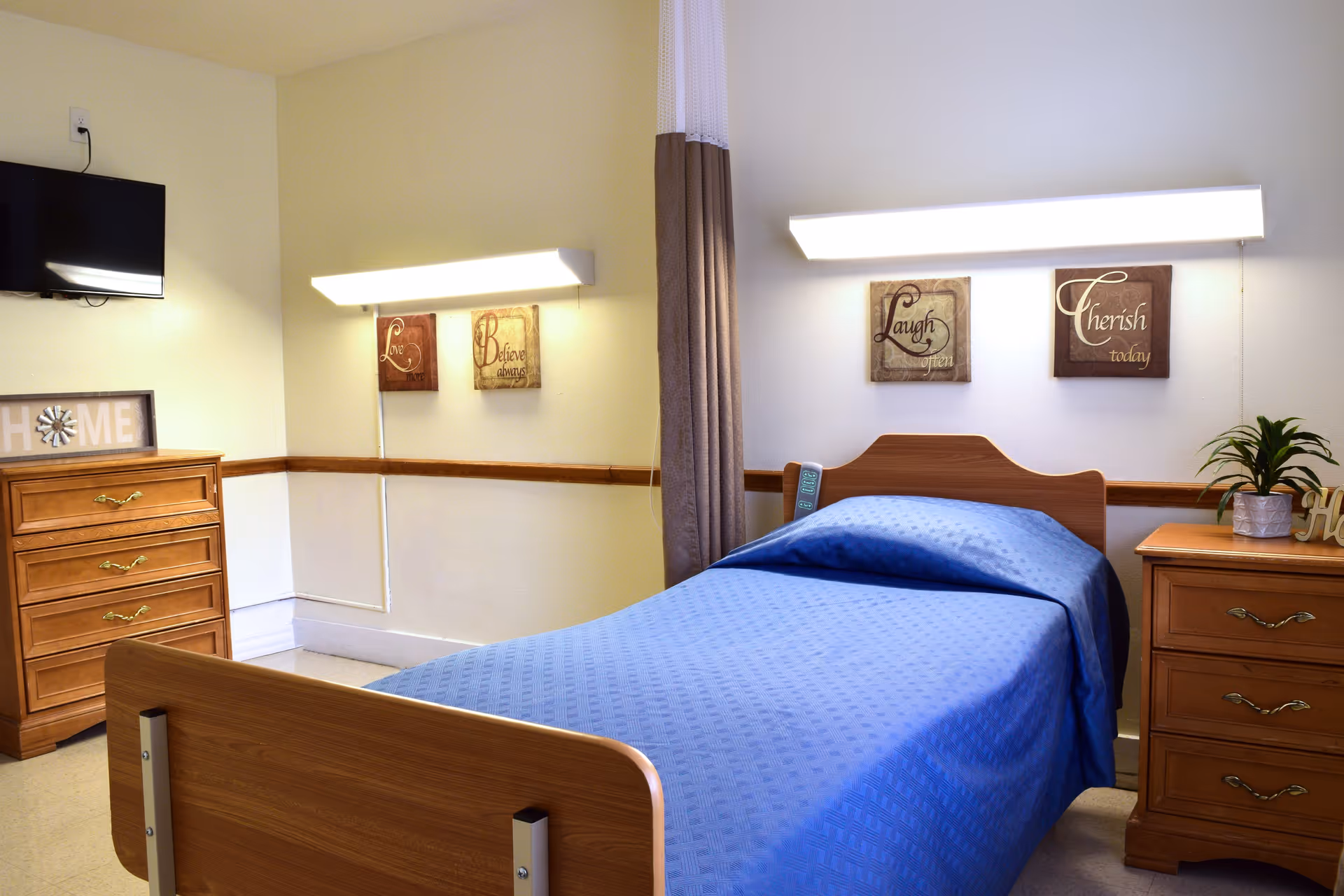A single bed with a blue blanket in a nursing facility room. The room has light yellow walls with wooden trim, two wooden dressers on either side of the bed, a small potted plant on one dresser, a wall-mounted TV, and four decorative wall plaques with the words 'Love', 'Believe always', 'Laugh often', and 'Cherish today'. There are two long wall lights above the bed and a privacy curtain partially visible.