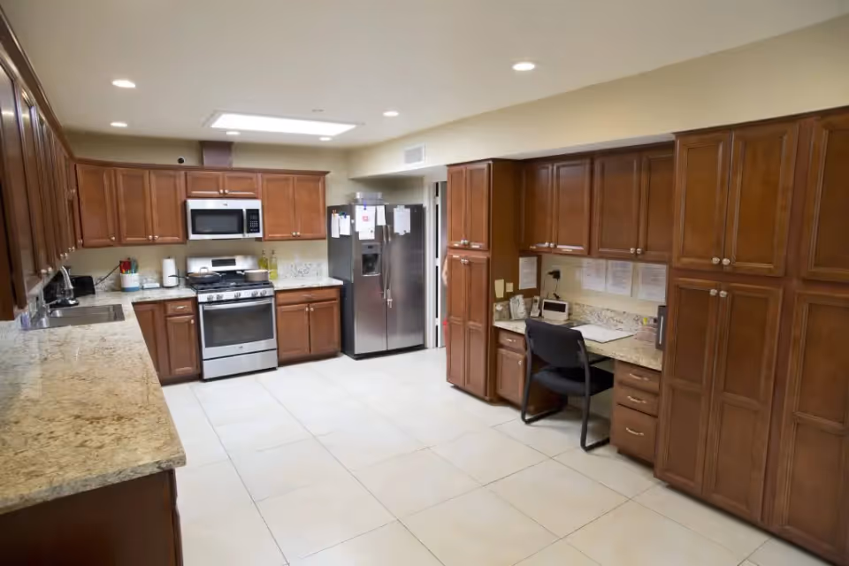 A spacious kitchen with wooden cabinets, granite countertops, a stainless steel refrigerator, stove, and microwave. There is a small desk area with a chair and papers on the counter. The floor is tiled with large light-colored tiles and the ceiling has recessed lighting and a skylight.